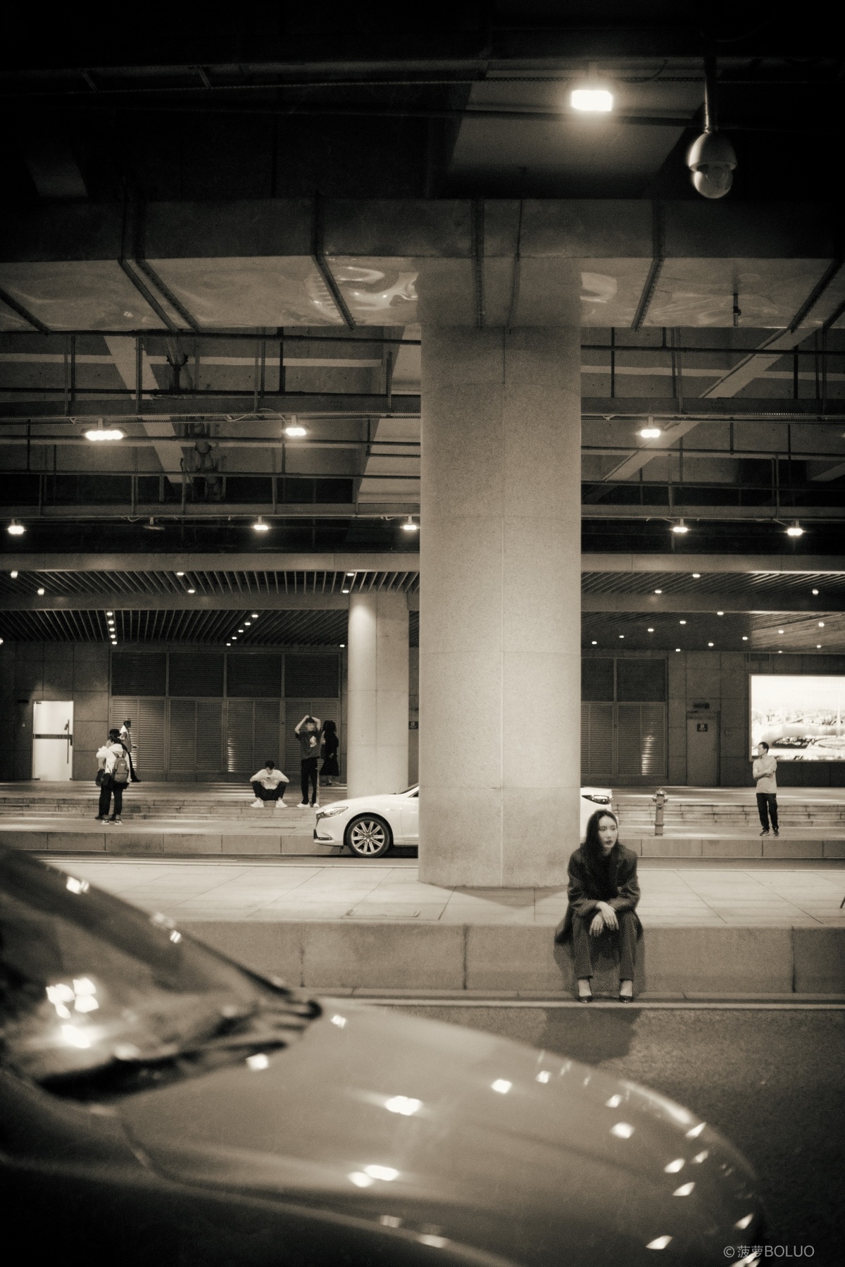 一张黑白照片 夜晚的停车场 街上有辆车 地上坐着一个人。