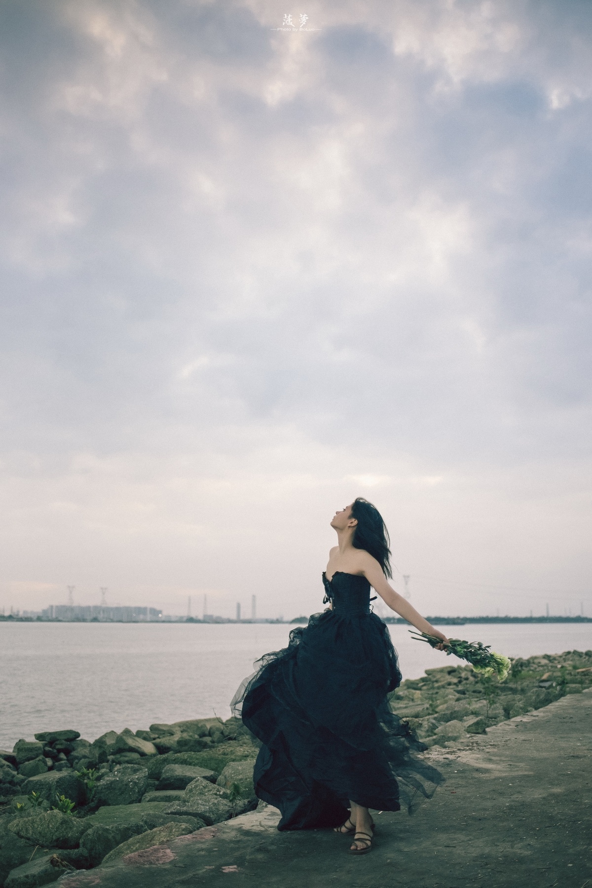 一位穿着黑色长裙的女性站在水体岸边岩石上