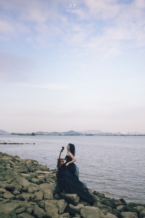 一位穿着连衣裙的女子坐在岩石上弹吉他 岩石位于水域的岸边。