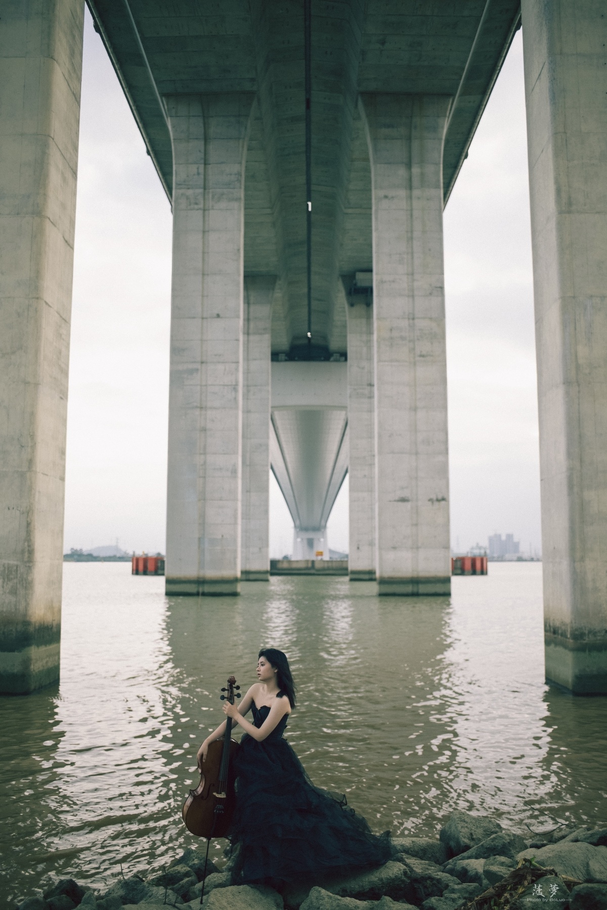 一个穿着连衣裙的女子坐在桥下方的岩石上弹奏小提琴