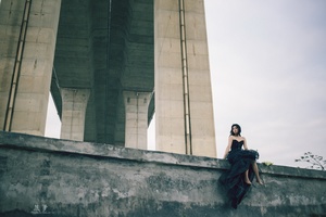 一个穿着黑色连衣裙的女人坐在桥上的一堵墙边 背景是一座建筑。