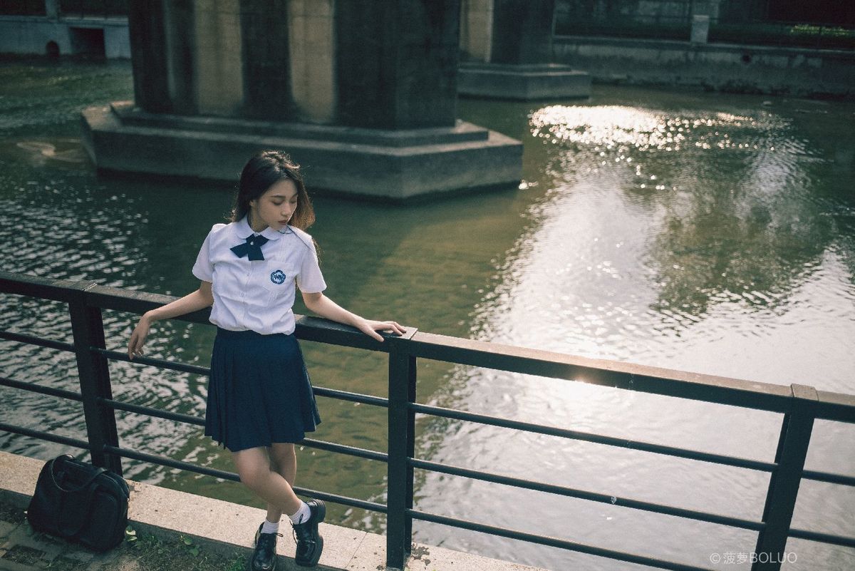 一个穿着校服的年轻女孩站在一座桥上 桥横跨在一条河上 手里拿着一个箱子。