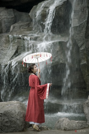 一位穿着红色连衣裙的小女孩手持雨伞站在瀑布前
