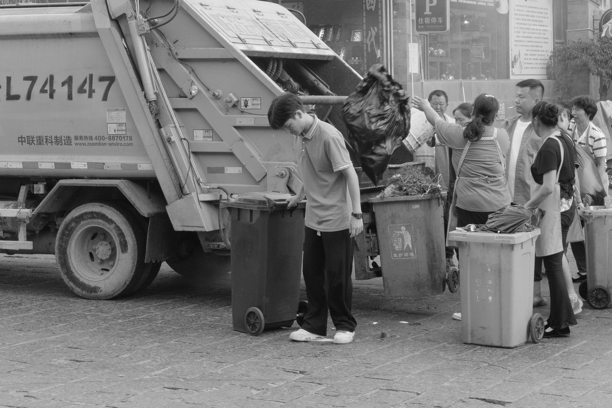 一张黑白照片 一个男子和一个男孩站在一辆正在向一些垃圾桶倒垃圾的卡车旁边。