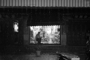 一张黑白照片 夜晚的街道 一个男人站在商店外面 自行车停在商店窗户前 一个女人带着伞。