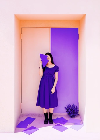 一位穿着紫色裙子的女士 手持一张纸在紫色的墙前。