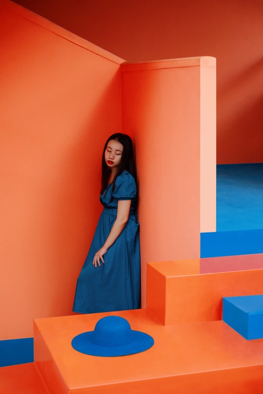 一位穿蓝色连衣裙和帽子站在彩色橙色墙壁前的女人