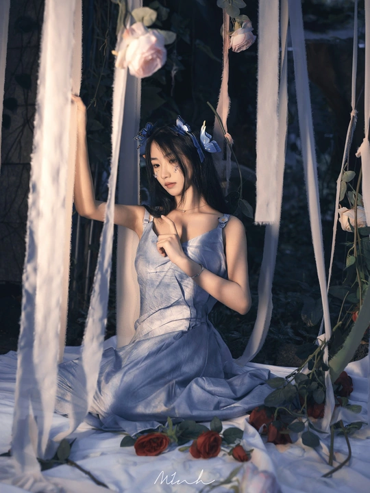 一个穿着蓝色连衣裙的年轻女子坐在用白色玫瑰花编成的秋千上。