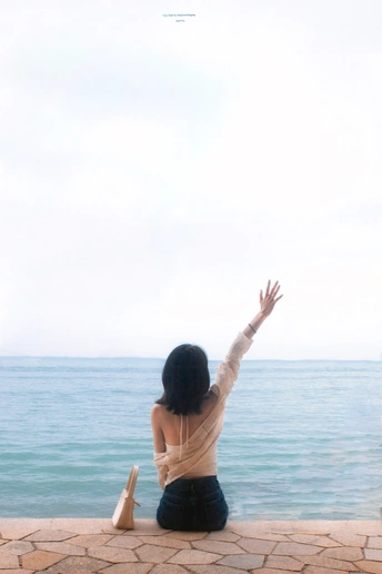 一位女子坐在墙上 手臂伸展 面向阳光下的海洋或海景。