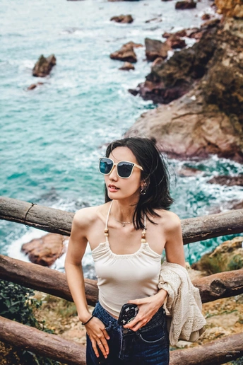 一个戴太阳镜和照相机的年轻女子站在一个俯瞰海洋和海岸线的岩石观景台上。