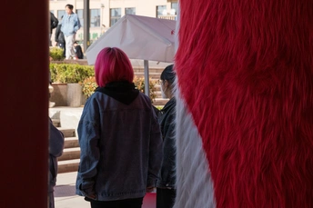 一位戴粉红色头发和红色假发的女士站在一把雨伞下 其他人经过。