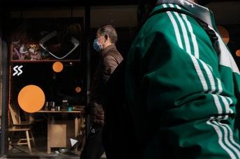 一个人穿着一件绿色夹克和戴着口罩走在街上 街对面是一家商店。