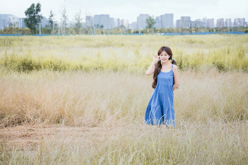 一个穿着蓝色连衣裙的年轻女子站在一片高草地上 正在用手机讲话。