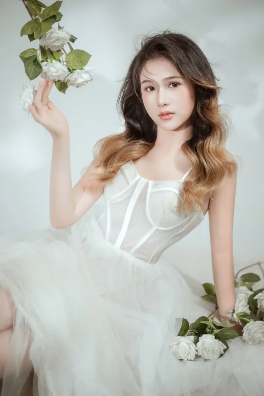 美丽年轻的女子穿着白色连衣裙 手持白色玫瑰 在白色背景上为肖像摆姿势的照片。