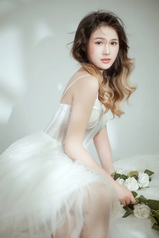 美丽年轻的女子穿着白色连衣裙 在白色背景上摆姿势 周围有白色玫瑰花。