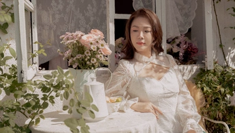 一个穿着白色连衣裙的年轻女子坐在窗前的一把椅子上 桌子上放着一束花。