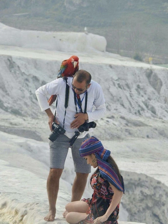 一个人肩膀上扛着相机 一个人肩膀上扛着鹦鹉 他们站在海滩上靠近一些人。