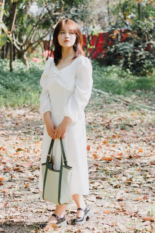 一个穿着白色连衣裙的女人拿着一个绿色手提包站在树叶里
