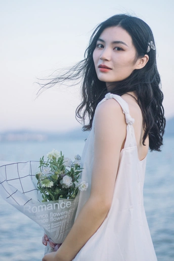 一位身穿白色连衣裙的年轻女子在水体附近手持一束鲜花的肖像