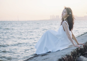 坐在海边或海边的墙上 穿着白色连衣裙的美丽年轻女子