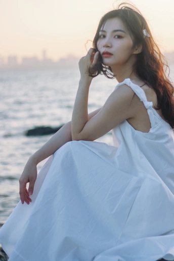 一个穿着白色连衣裙的美丽年轻女子坐在海滩水边的岩石上