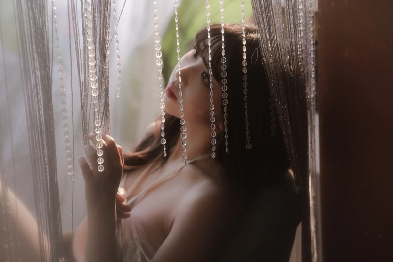 一个美丽的年轻女子站在窗前  behind 一层薄纱窗帘后面。