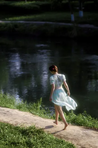 一位穿着白色连衣裙的年轻女子沿着池塘或河流附近的小径走