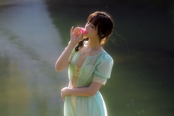 一位穿着绿色连衣裙的年轻女子从一朵花中喝水 站在湖或河边。