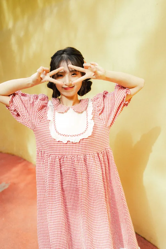 一个小女孩穿着红白相间的格子裙 把鼻子贴在耳朵上 摆出pose。