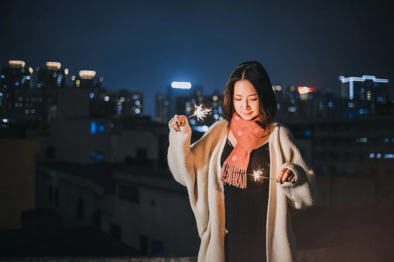 一个穿着外套的年轻女子手持点燃的焰火站在屋顶俯瞰城市夜景