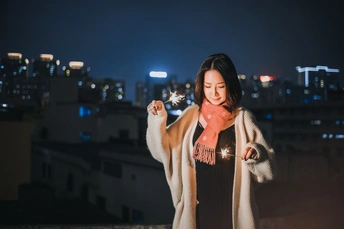 一个穿着外套的年轻女子手持点燃的焰火站在屋顶俯瞰城市夜景