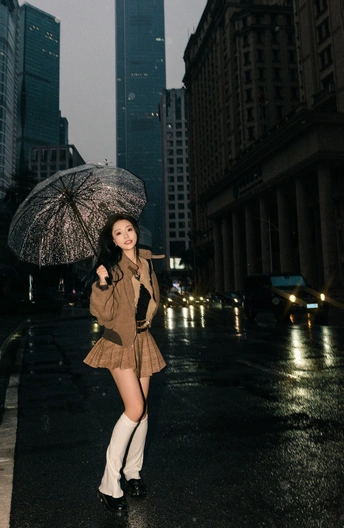 一个穿靴子和带着雨伞的年轻女子站在城市街道上 而另一个年轻女子正在街上走。