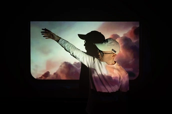 一个戴帽子的人在窗户前站立 双手伸开 一个女性站在他身后。