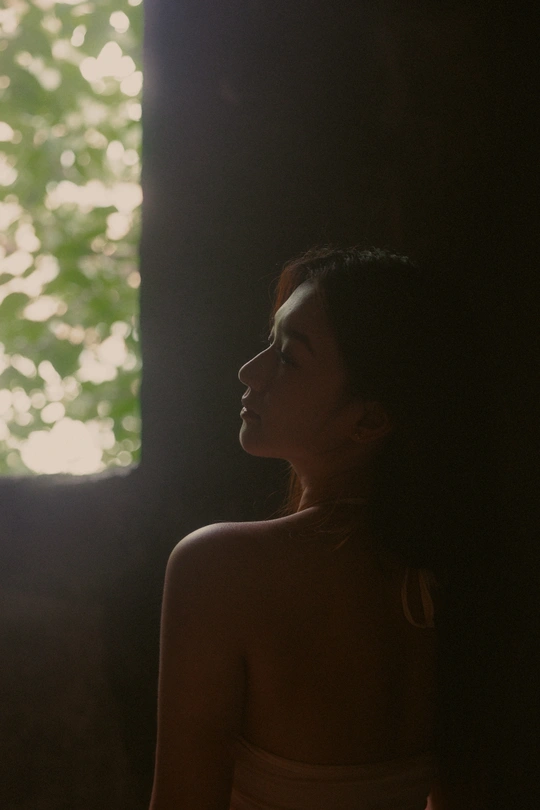 一个穿着连衣裙的年轻女子站在一个光线昏暗的房间里 背景中有一棵树。