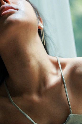 一位年轻女子穿着一件白色吊带衫 正通过窗户凝视着 同时拿着一部白色手机贴在耳边。