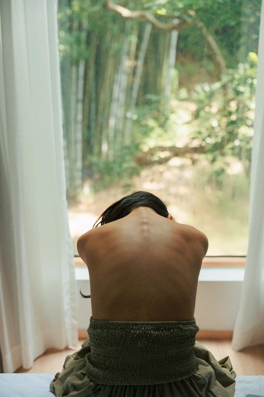 一个女性坐在床上 正在透过窗户向外看。