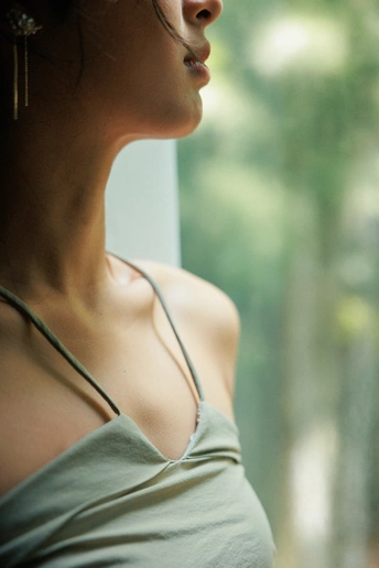 一位穿着背心的年轻女子正在窗边凝视着窗外的树木。