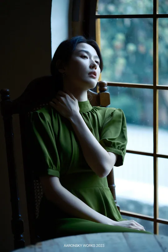 一个穿着绿色连衣裙的年轻女子坐在桌子旁边 朝窗外望去。