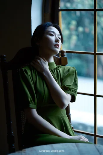 一个穿着绿色连衣裙的年轻女子坐在桌子旁边 朝窗外望去。