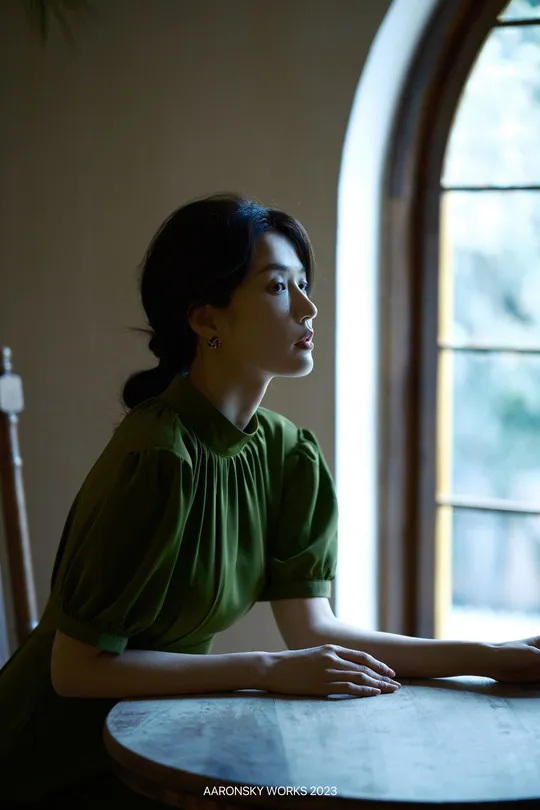 一位穿着绿色衣服的年轻女子坐在桌子旁 凝视着窗外。