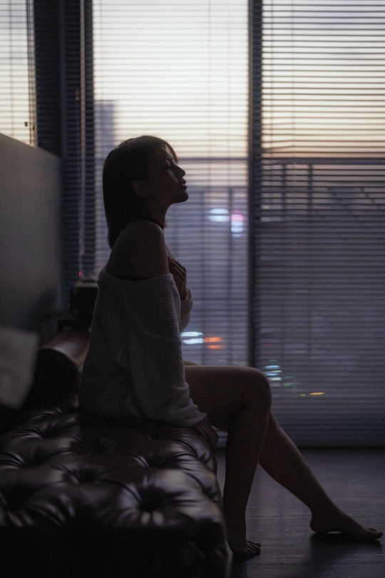 一个年轻女子坐在一个黑暗的房间里 房间里有窗户和电视。