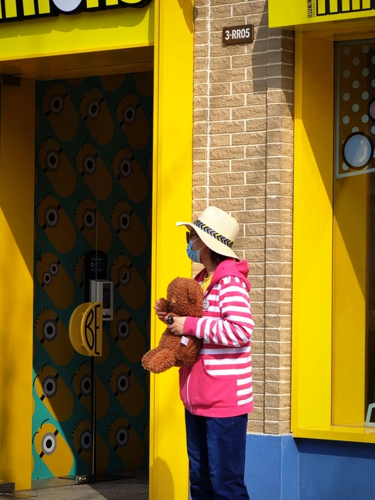 一位戴着粉红色帽子的小女孩站在一家黄色商店前 手里拿着一只棕色泰迪熊。