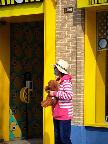 一位戴着粉红色帽子的小女孩站在一家黄色商店前 手里拿着一只棕色泰迪熊。