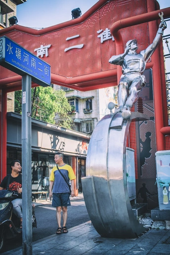 摩托车上的巨大男性雕像站在一个城市的入口处 周围有建筑物和行人。