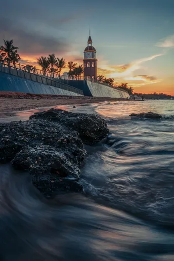 太阳正在落下一片水域 这片水域附近有一座灯塔和岩石。