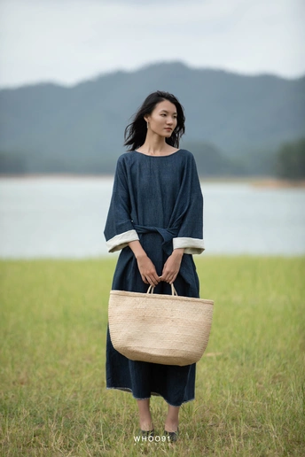 一位穿着蓝色连衣裙的年轻女子 手提篮子 穿过一片靠近水域的田野 拿着一个袋子。