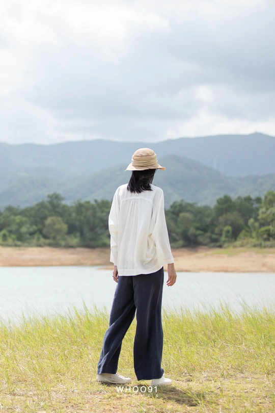 一位穿着白色衬衫和帽子的年轻女子站在一个俯瞰湖泊和山脉的田野上。