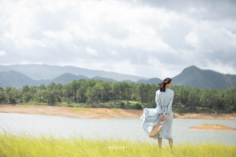 一位穿着蓝色连衣裙的年轻女子站在高草丛中 背景是湖泊和山脉。
