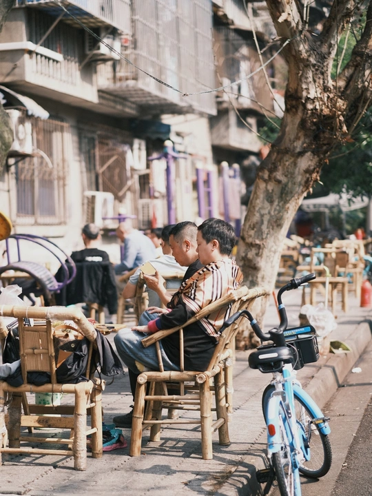 一个年轻男孩坐在人行道上的一把木椅上 与此同时 一个男子坐在街边的一张桌子旁 桌子上有一辆自行车。