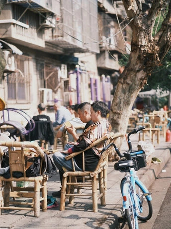 一个年轻男孩坐在人行道上的一把木椅上 与此同时 一个男子坐在街边的一张桌子旁 桌子上有一辆自行车。